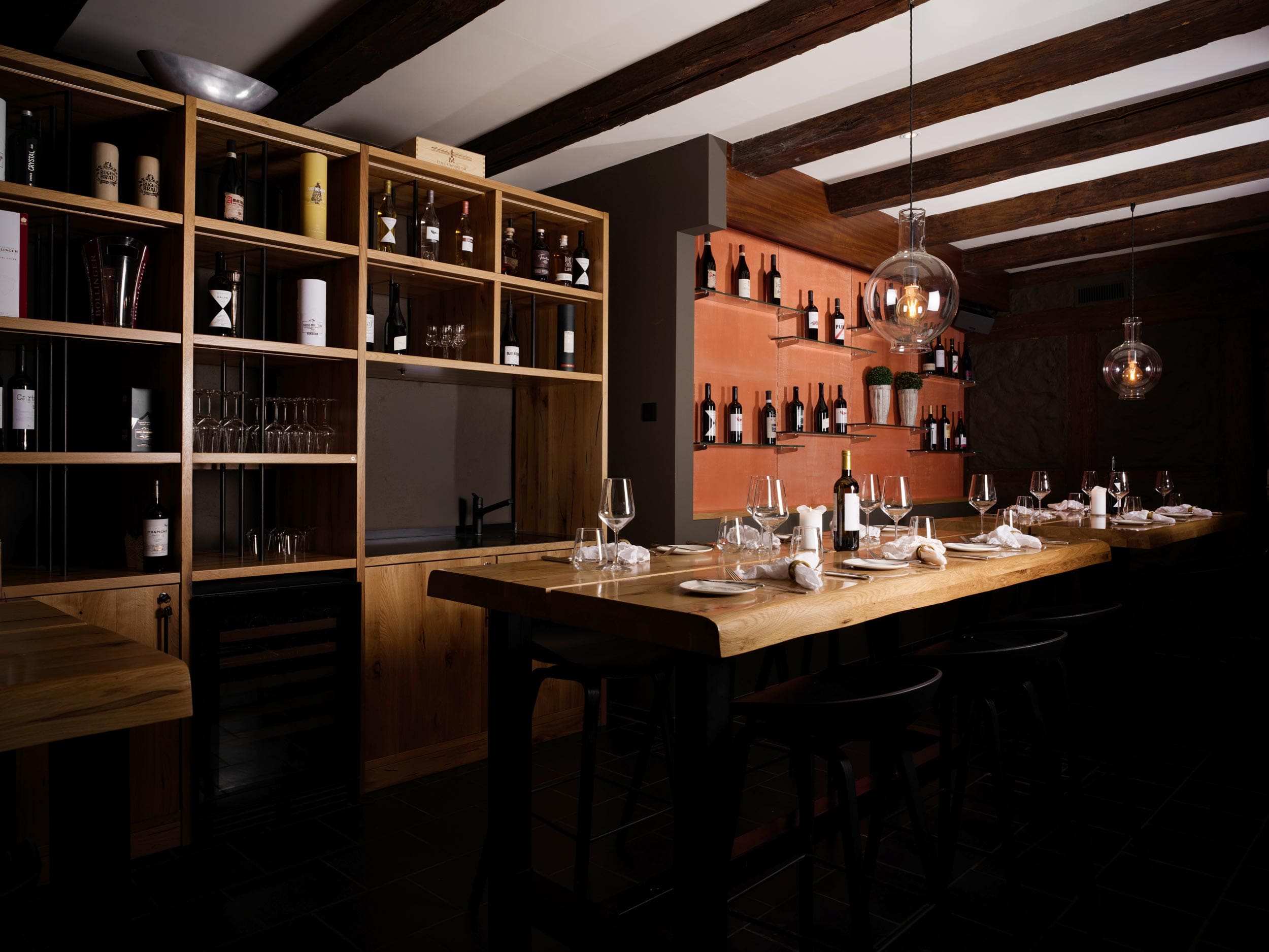 Un bar à vin moderne, faiblement éclairé, avec des tables en bois, des étagères de bouteilles de vin, des lampes en verre suspendues et des tables soigneusement dressées avec des verres à vin, des serviettes et des couverts, créant une atmosphère confortable et élégante.