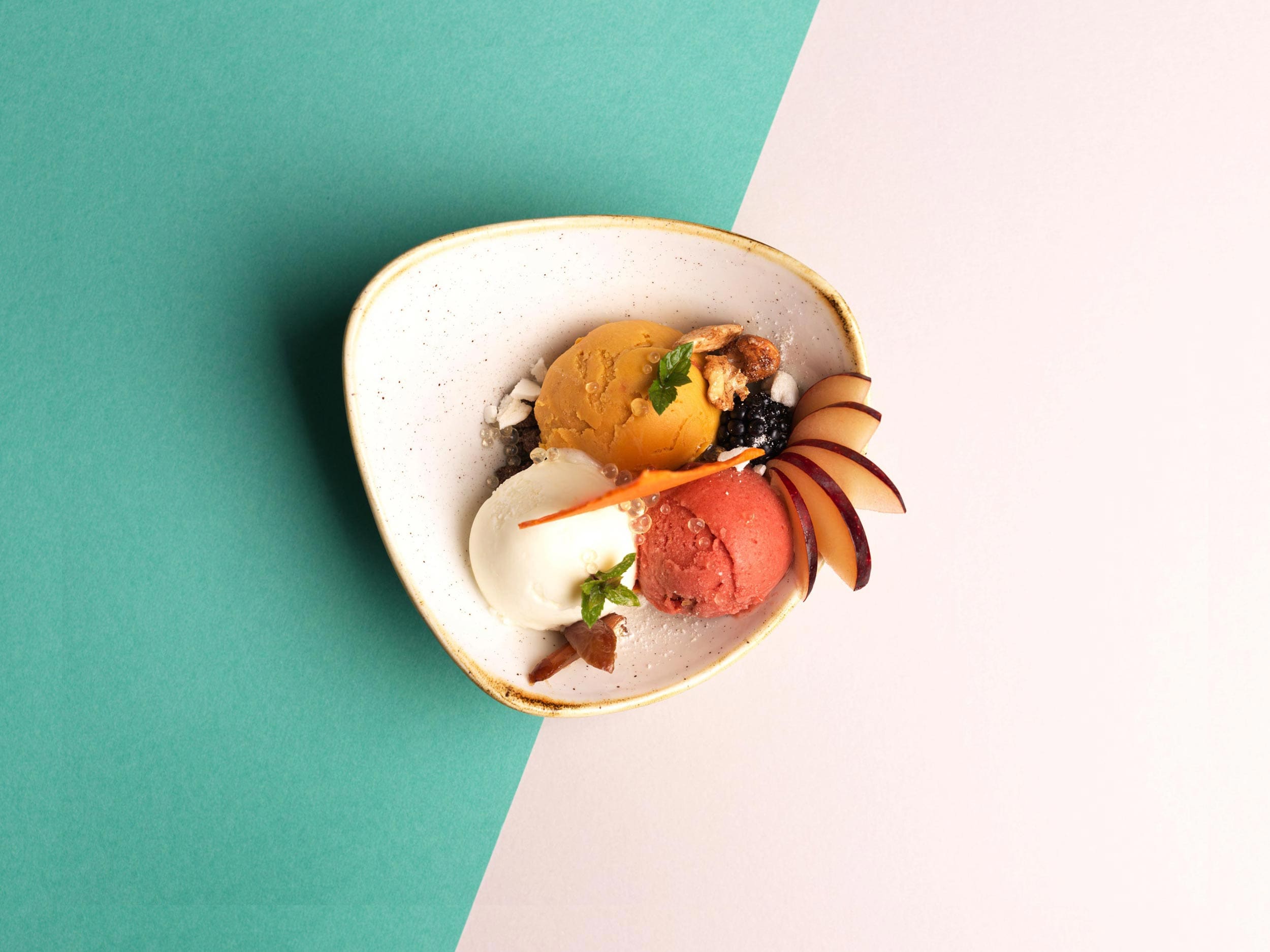 Un bol avec trois boules de glace (jaune, blanche et rose) garnies de menthe, de noix et de tranches de prune, placé sur un fond vert et blanc partagé.