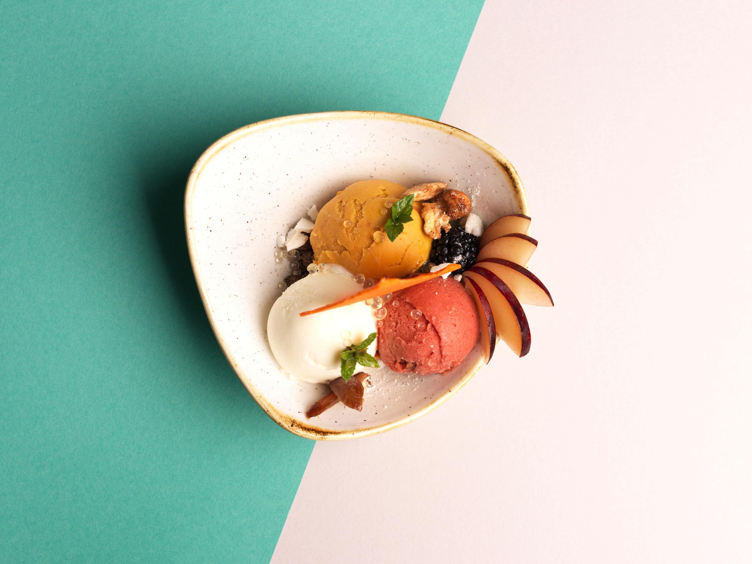Eine Schale mit drei Kugeln farbenfrohen Sorbet - weiß, gelb und rot - garniert mit Pflaumenscheiben, Minze, Nüssen und Krümeln, platziert auf einem geteilten grünen und gebrochen weißen Hintergrund.