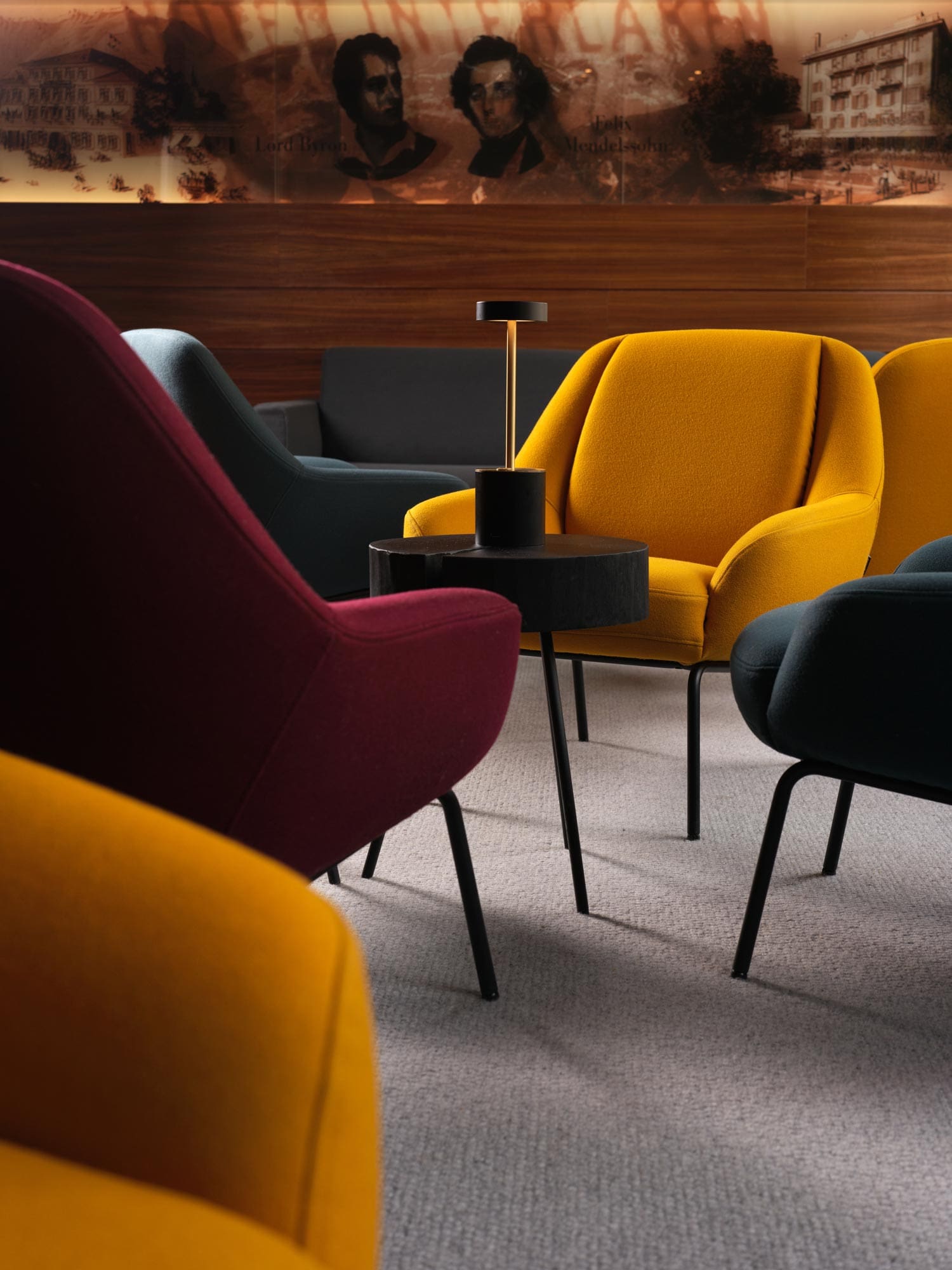 Salon moderne avec des fauteuils jaune moutarde, violet foncé et noir autour d'une petite table ronde avec une lampe noire élégante ; une peinture murale avec des portraits décore le mur en bois à l'arrière-plan.