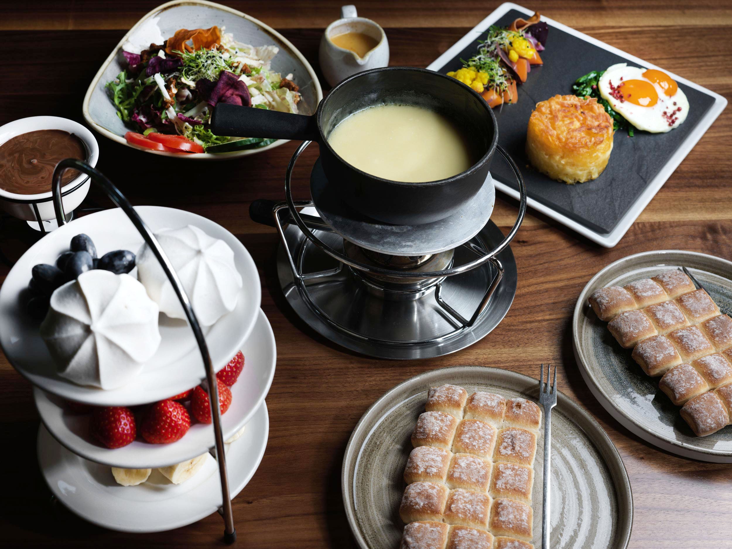 Une table avec un service à fondue au fromage, une salade, des légumes grillés avec un œuf, des rösti, des fruits frais, des gaufres, des meringues, de la sauce au chocolat et du café sur une surface en bois.