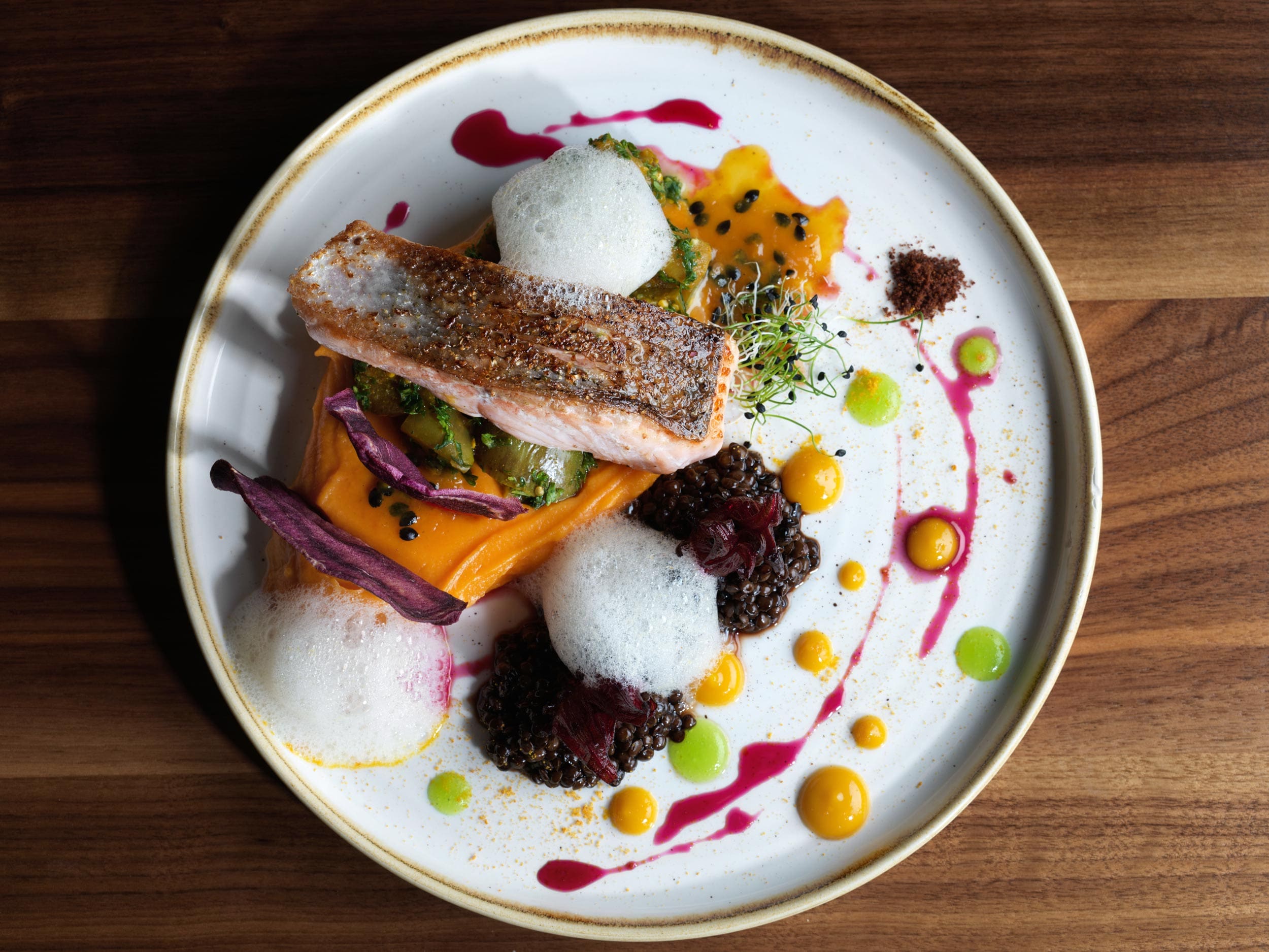 Le 5th Floor Jnterlaken propose un plat gastronomique composé d'un filet de saumon saisi sur une purée d'orange vibrante, avec une sauce blanche mousseuse, des lentilles noires, des sauces colorées et des garnitures disposées avec art sur une assiette blanche.