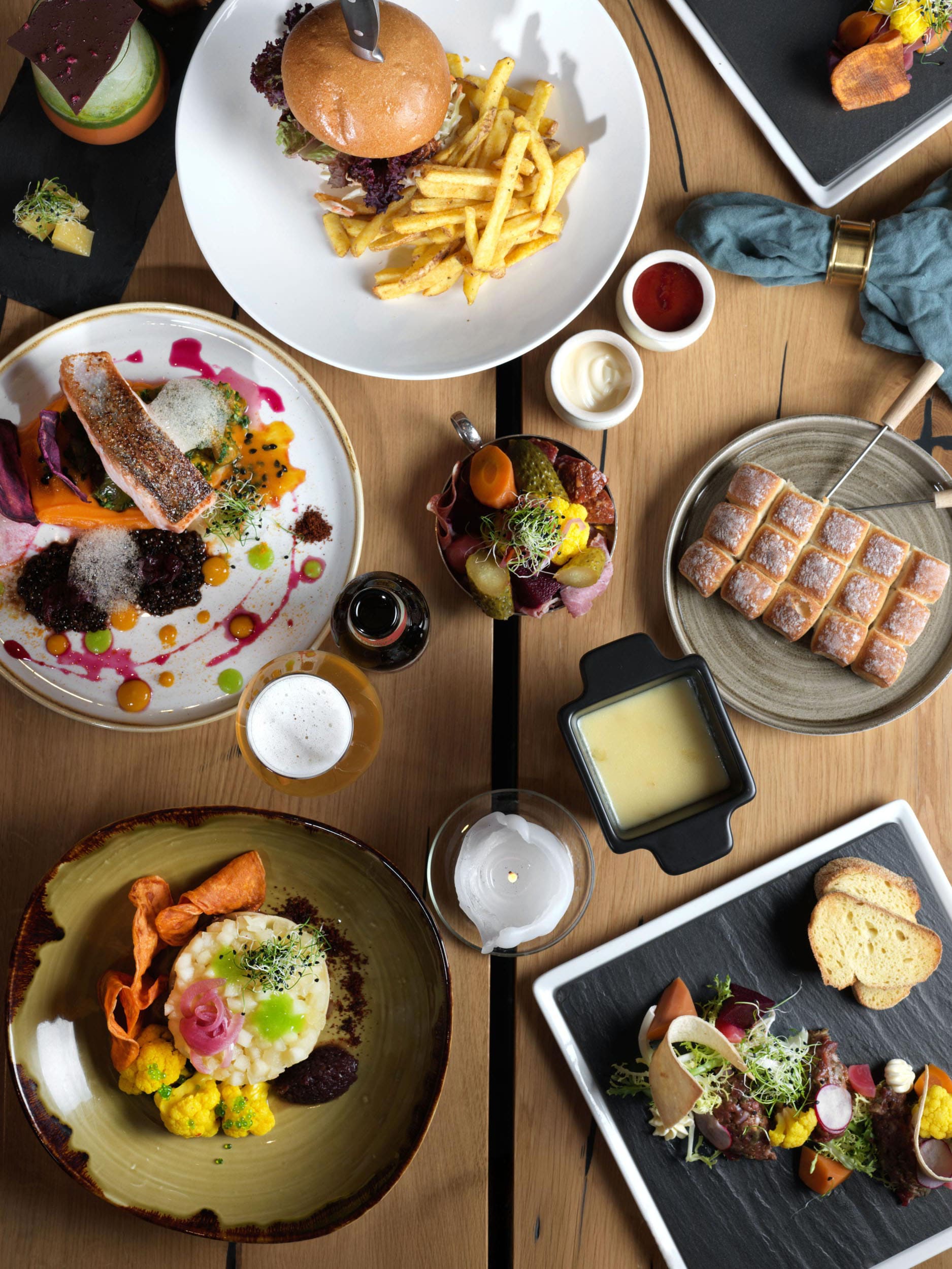 Vue de haut en bas d'une table en bois garnie de plats gastronomiques assortis, dont un hamburger avec des frites, du saumon avec des légumes, une fondue au fromage, une salade, du pain, des sauces et des boissons, le tout disposé de manière attrayante.