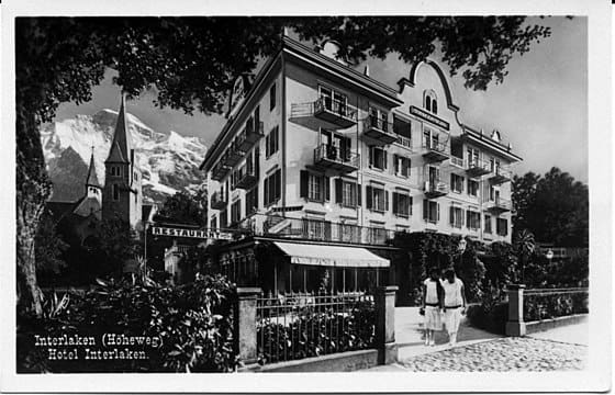 Photo en noir et blanc de l'hôtel Interlaken en Suisse, avec des personnes marchant devant, une église avec un haut clocher à proximité et des montagnes enneigées en arrière-plan.