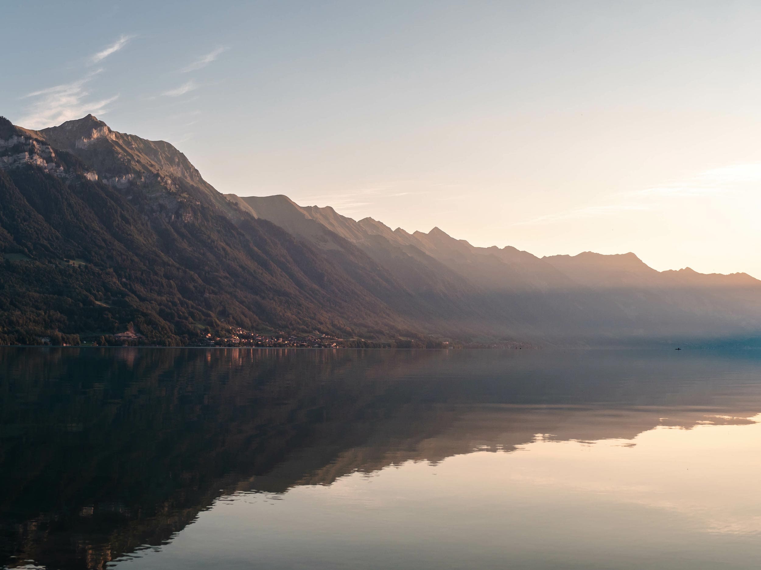 Lac calme avec des reflets clairs des montagnes escarpées au lever ou au coucher du soleil, une lumière douce projetant des ombres douces sur les pentes, et un ciel pâle au-dessus de l'eau tranquille.
