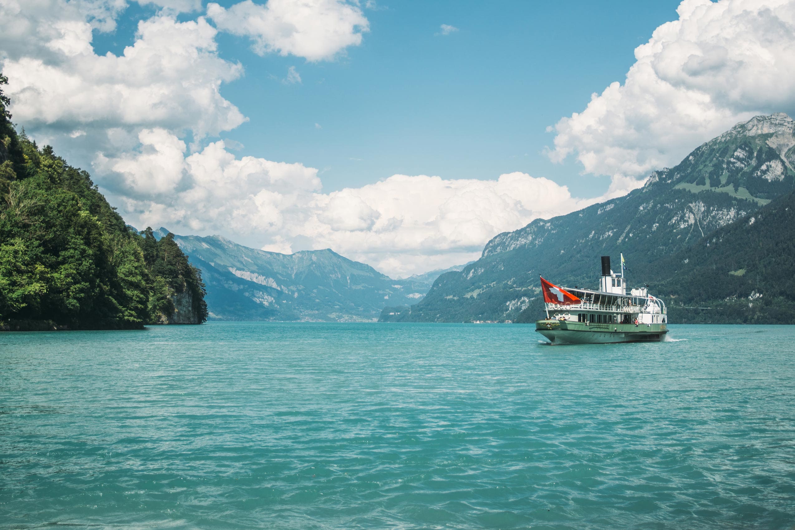 Un bateau de passagers blanc avec un drapeau rouge navigue sur un lac turquoise entouré de montagnes vertes et de pics lointains sous un ciel partiellement nuageux.
