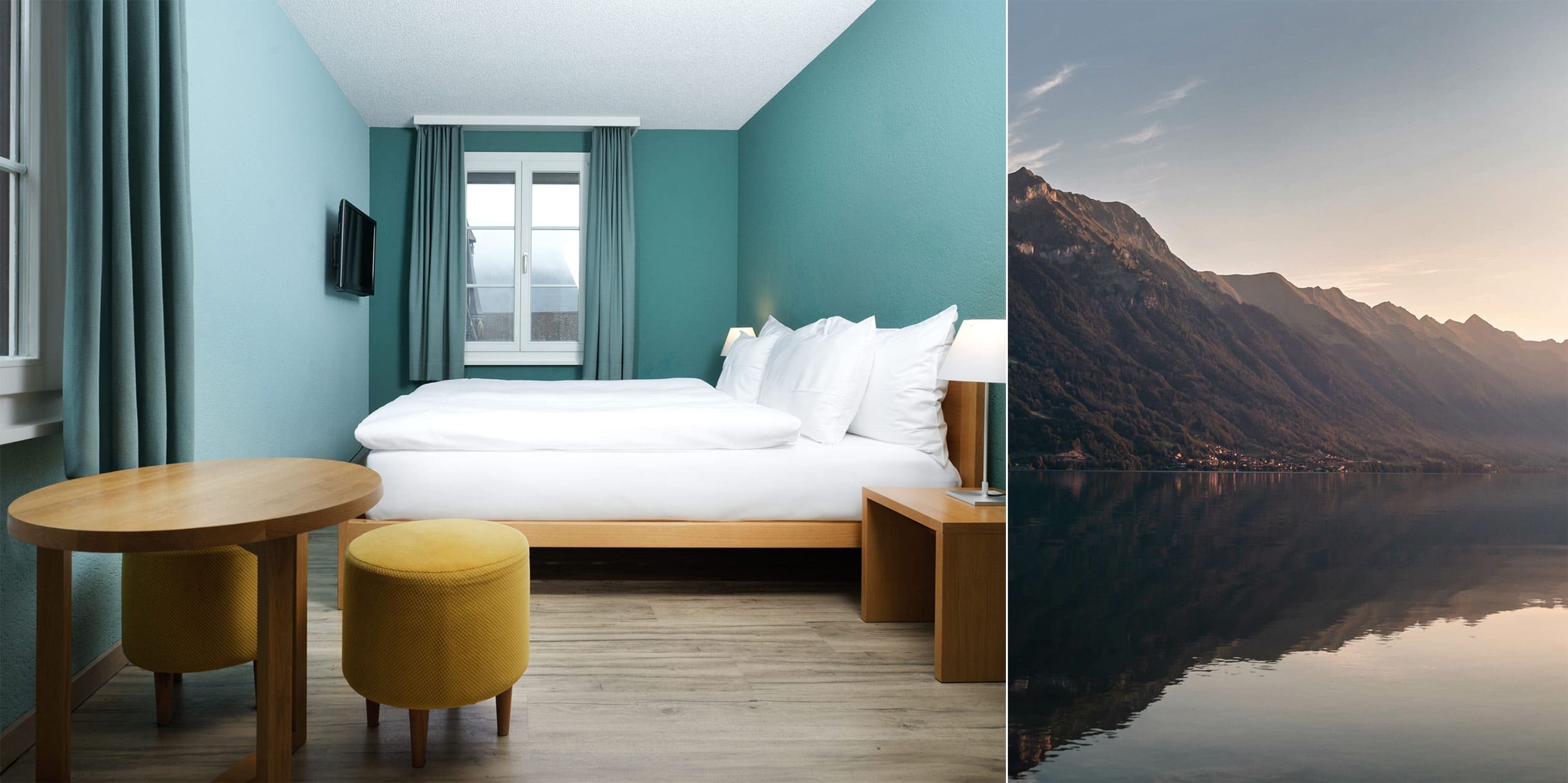 Ein modernes Hotelzimmer mit tealfarbenen Wänden, einem großen Bett mit weißer Bettwäsche, einem runden Holztisch, zwei gelben Hockern und einem Fenster; daneben ein ruhiger See mit Bergen im Sonnenuntergang.